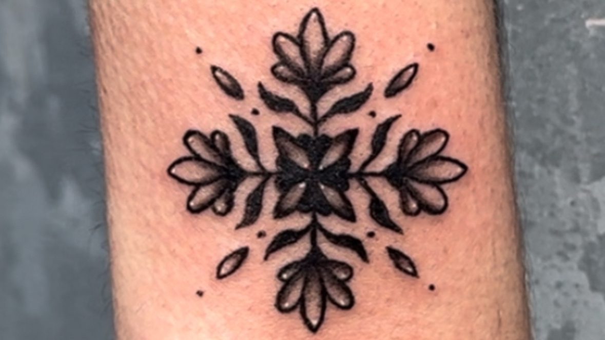 A detailed black tattoo of a snowflake design featuring floral elements, placed on the forearm.