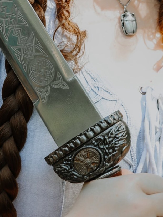 Close-up image of a hand holding a decorative sword with intricate engravings on the blade and a detailed hilt, set against a backdrop of braided hair.