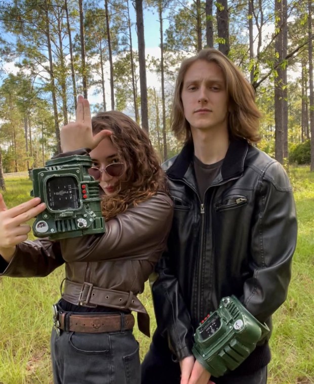 A woman in a brown leather jacket and sunglasses stands striking a pose with a green device on her wrist, while a man in a black leather jacket stands beside her with a serious expression. They are surrounded by tall trees in a grassy area.