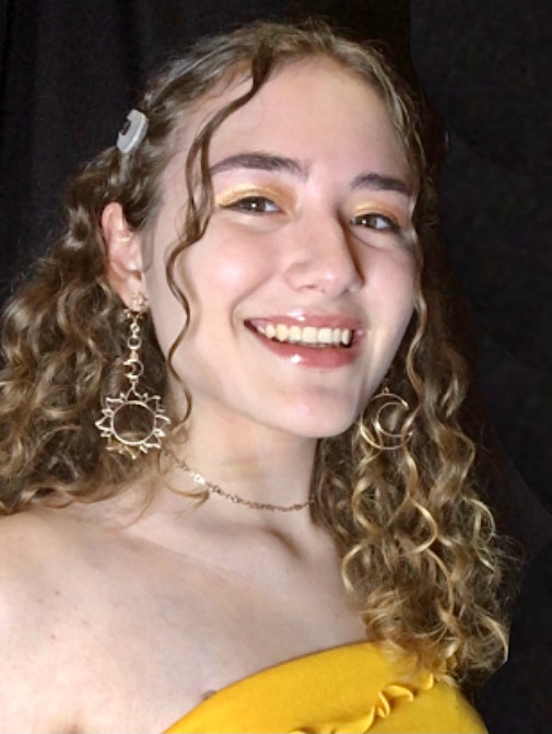 A young woman with curly hair and a bright smile, wearing a yellow top and decorative earrings, poses for a photo against a dark background.
