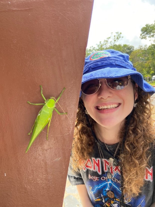 A smiling person wearing a blue hat and sunglasses stands beside a large green insect on a post.