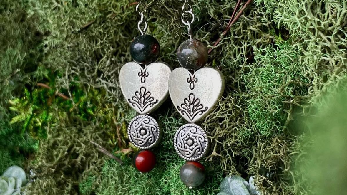 A pair of handmade earrings featuring silver hearts and decorative beads, displayed on a bed of green moss.