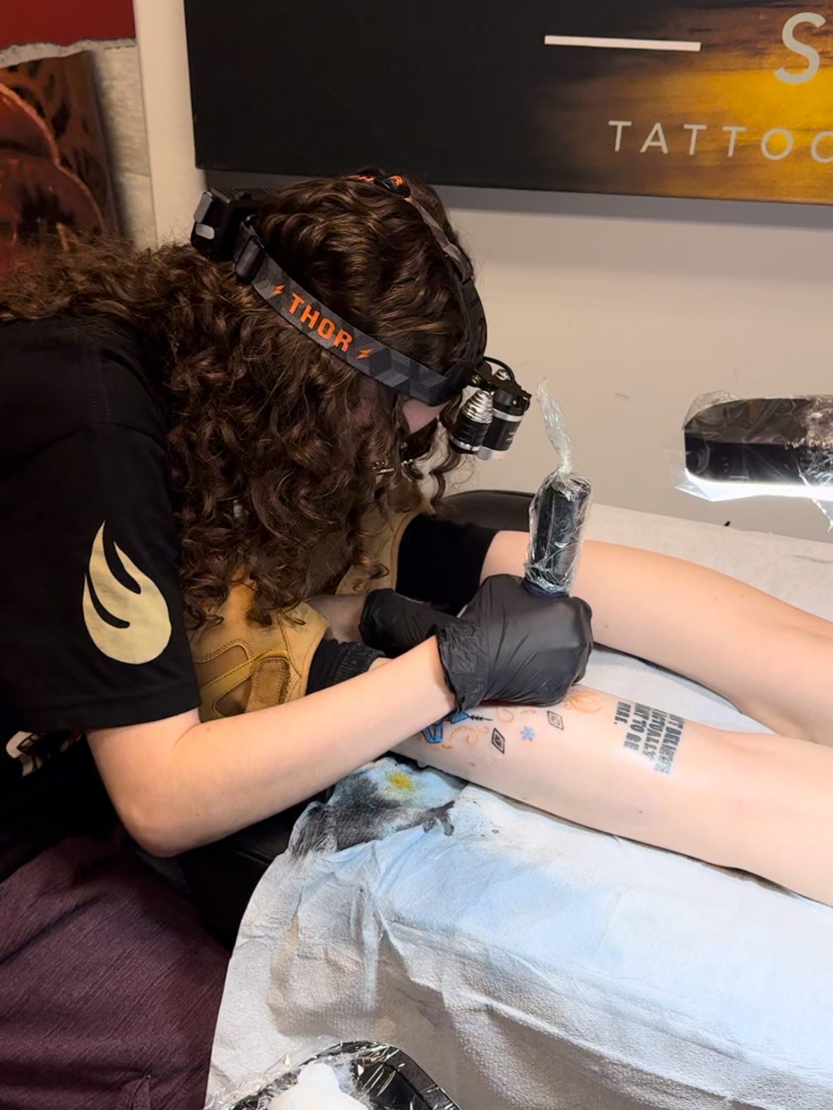 A tattoo artist with curly hair applying a colorful tattoo on a person's thigh while using a tattoo machine, with a studio background.
