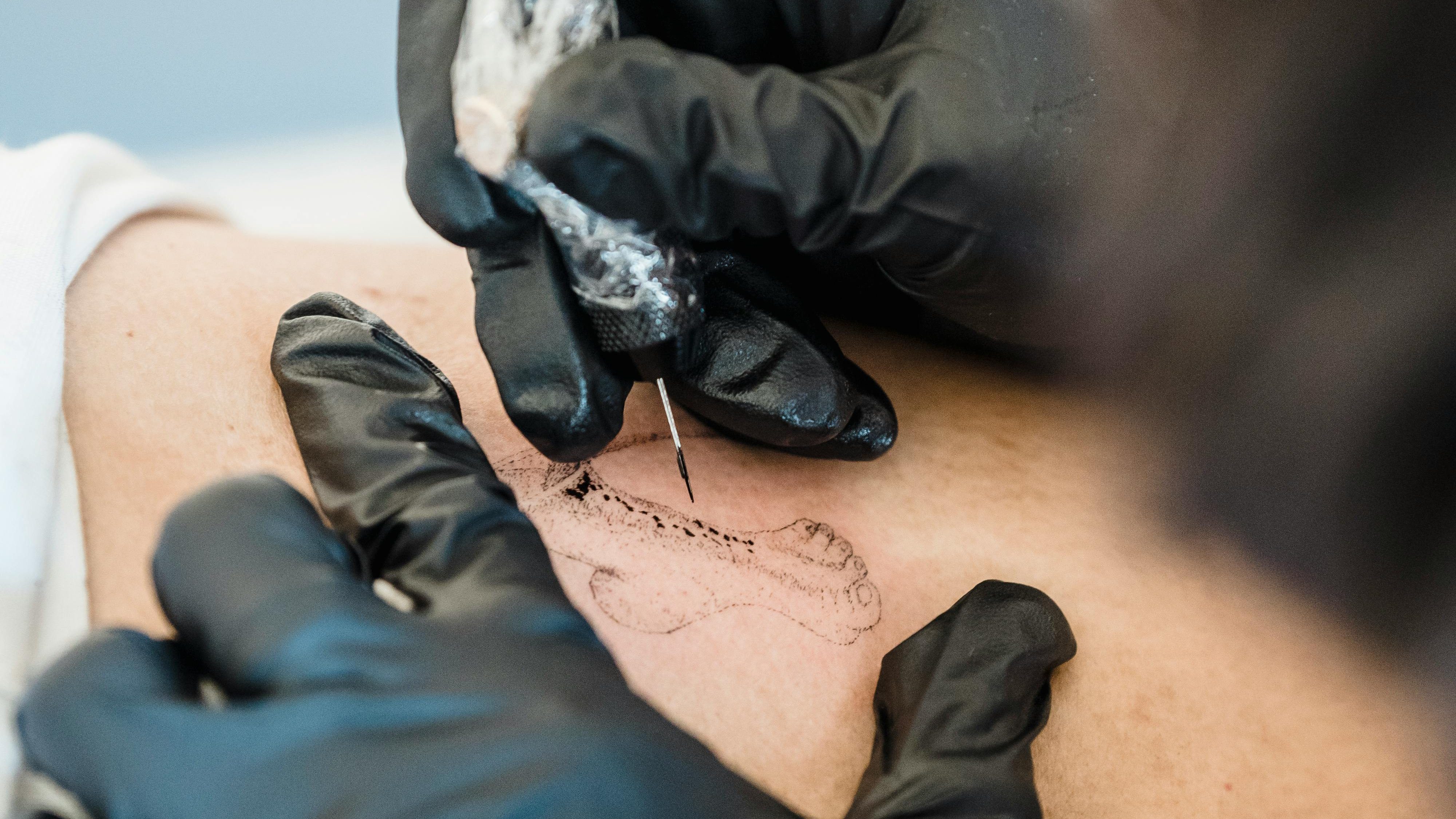 A close-up of a tattoo artist's hand in black gloves using a needle to tattoo a foot design on skin.