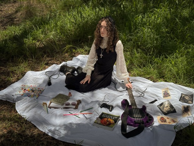 A young woman sitting on a white blanket in a grassy area, wearing a black dress and a long-sleeve white top. She has curly hair and is surrounded by various items including a purple bass guitar, art supplies, books, and images, reflecting her artistic pursuits.
