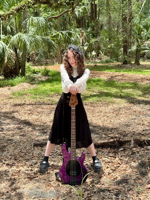 A young woman with curly hair standing in a forest, wearing a black dress and a white lace blouse, holding a purple bass guitar in front of her.
