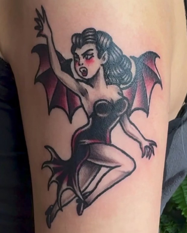 A colorful tattoo of a pin-up style vampire woman with bat wings, wearing a black dress and striking a dynamic pose.