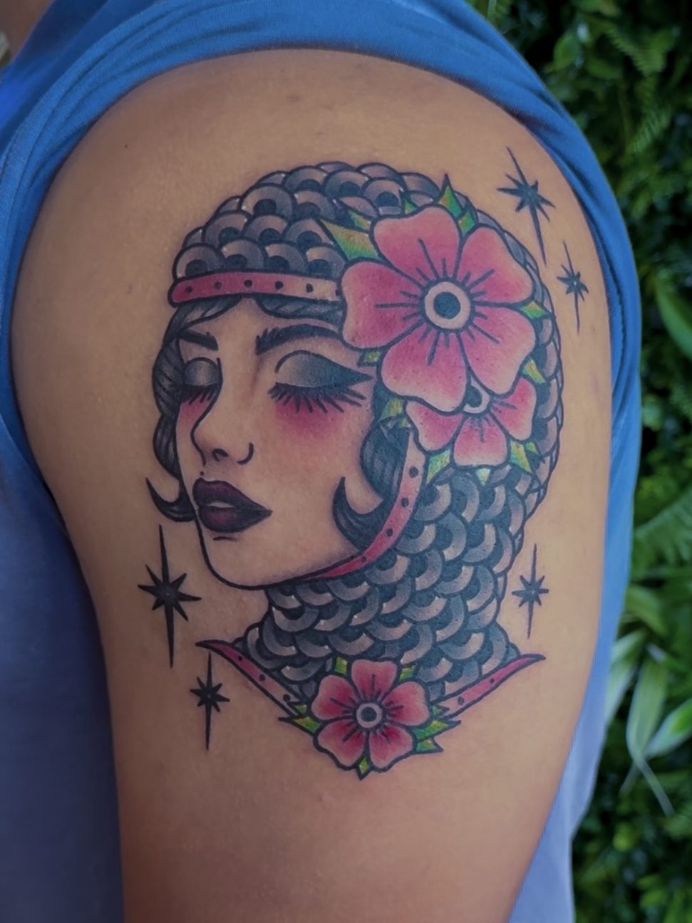 A close-up of a colorful tattoo depicting a woman with braided hair, wearing a headpiece, and surrounded by flowers and stars, inked on a person's upper arm.