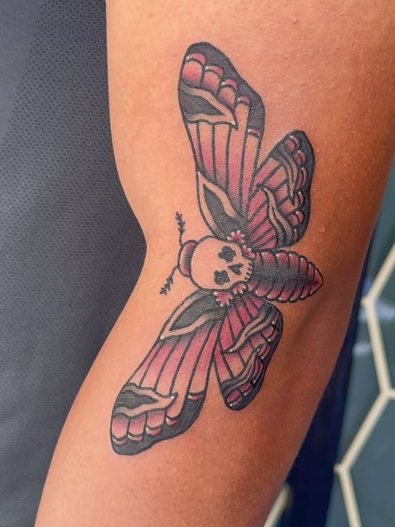 Tattoo of a moth with decorative patterns and a skull design on a person's forearm.