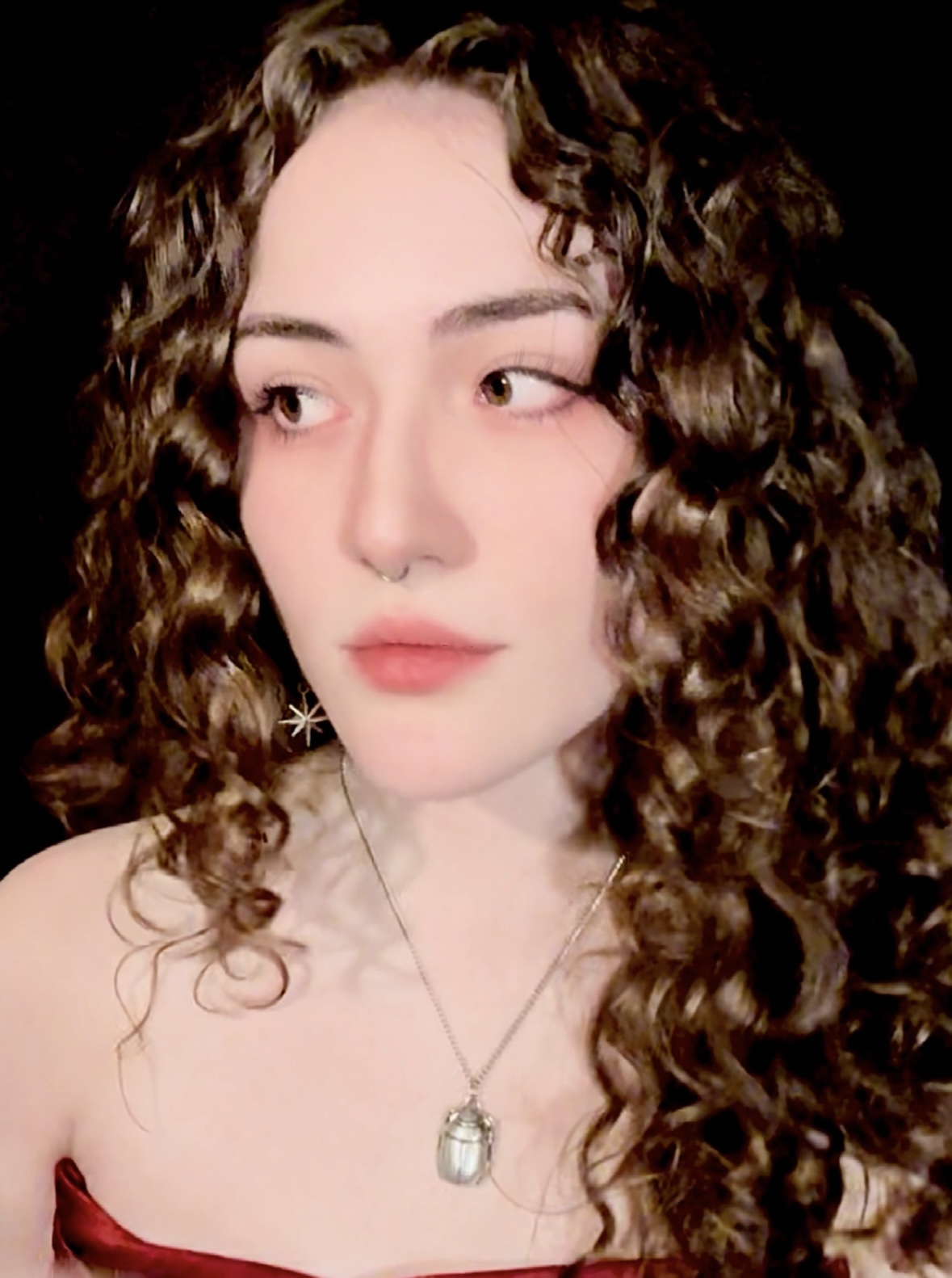 Close-up portrait of a person with curly hair, wearing a red top and silver jewelry, including a star-shaped earring and a pendant, against a dark background.